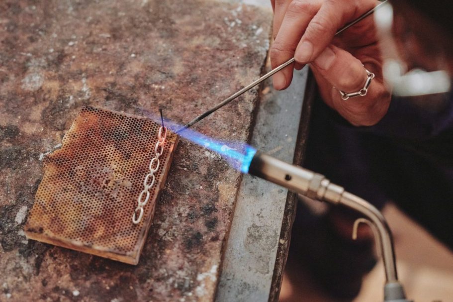 Spezialanfertigungen Eine Hand hält einen Brenner, der eine Kette auf einer Metallplatte erhitzt.