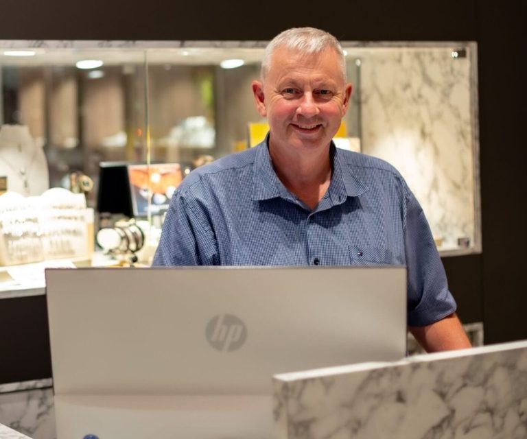 Inhaber Sven Born Mann am Schalter mit Laptop, lächelt freundlich. Schmuckauslage im Hintergrund.